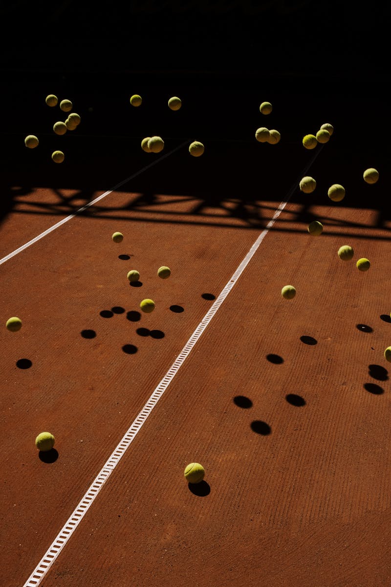 Tennis balls in mid-air on a clay court create a dynamic motion effect with dramatic shadows.