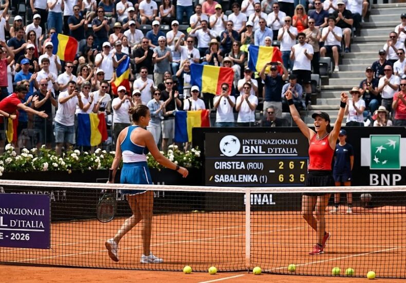 Two female tennis players on a red-clay court after a match, one with arms raised in victory as the crowd cheers with Romanian flags behind them.